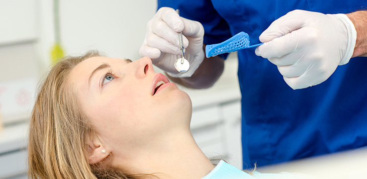 young adult woman laying back in a dentists chair while the dentist is holding a mirror and mouth guard over her face