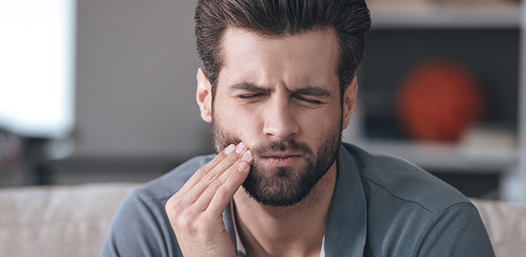 man with big hair and a hipster beard pushing on his cheek with his eyes closed as if he is in pain